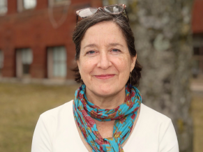 White woman with brown hair, blue eyes, smiling standing in front a tree and a building, wearing a printed scarf, white shirt, and glasses rest on head. 