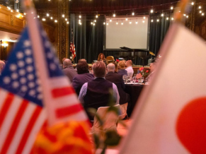 Japan and American flag out of focus. Man watches auditorium filled with people.